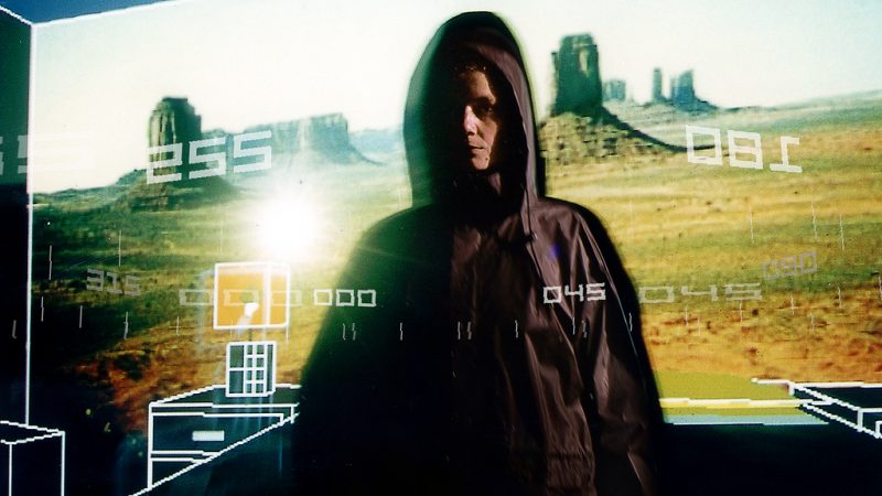 Promotional image for Desert Rain (1999). A white woman standing in a big parka coat. Behind them is a projector featuring a image of a desert. CGI numbers overlay the image.