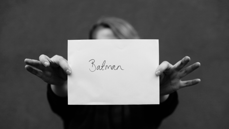 A young woman holding up a piece of paper with "Batman" written on it, covering her face.