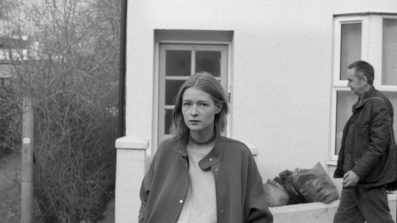 Black and white photo of a young woman with dark blond hair. She is outside a house and is wearing a bomber jacket.