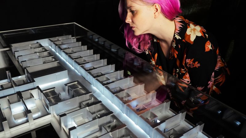 A woman with pink hair looks at a model of a hotel room made from metal