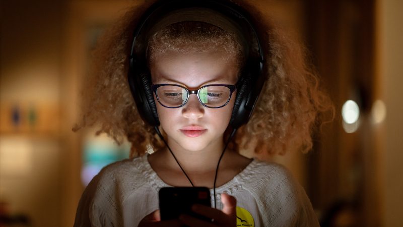 Close up of a child wearing glasses and large headphones, holding a mobile phone. The light from the screen glows on their face.