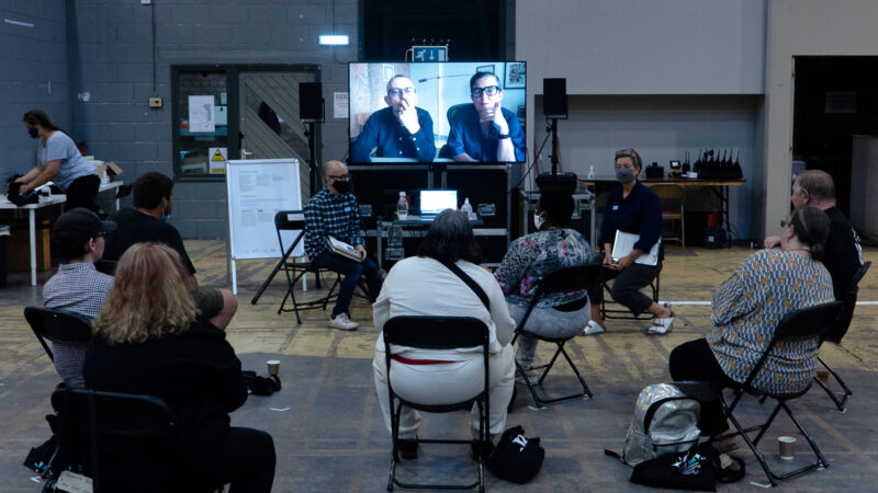 Seven Manchester Street Poem members are sitting in a large industrial space, looking at artist Nick Tandavanitj and Associate Artist Niki Woods. Artists Ju Row Farr and Matt Adams are visible on a large screen.