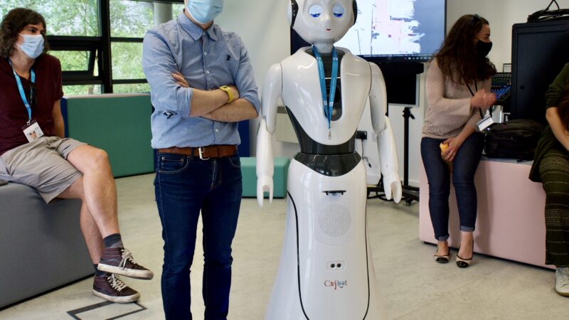 Blast Theory artist Nick, wearing a mask, hat, shirt and trousers, stands cross-armed next to a human-sized white robot with human facial features, arms and a circular body.