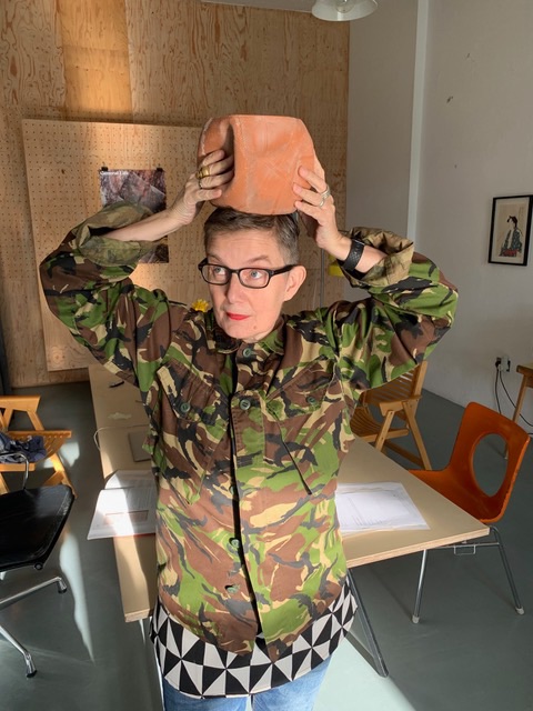 Artist Ju is wearing a camouflage jacket with a black and white abstract top underneath. She is holding a plant pot on top of her head and is staring off into the distance.