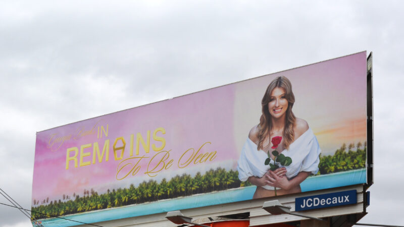 A billboard installed on top of a building depicts a smiling blonde woman holding a rose. Behind her is a pink and orange sunset, palm trees, and blue water. A gold logo read ‘Georgia Banks in Remains To Be Seen’