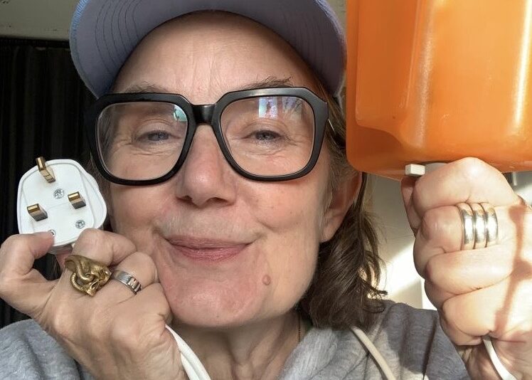 A smiling white women, wearing a cap and glasses. Holding an orange lamp in one hand and the plug socket in the other.